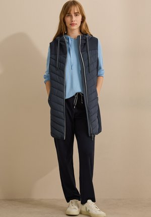 Gilet imbottito lungo blu navy con zip frontale e cappuccio, caratterizzato da cuciture a zigzag. Abbinato a una felpa blu chiaro, pantaloni scuri e sneakers bianche.