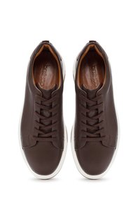 Zapatillas de cuero marrón con suelas blancas, que presentan detalles perforados y cordones tradicionales. El interior tiene un forro suave de color marrón claro.