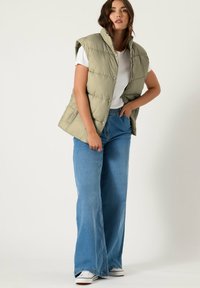 Kamizelka puchowa w jasnym oliwkowym kolorze z zapięciem na zamek błyskawiczny i zatrzaskami, połączona z szerokimi niebieskimi dżinsami i białymi trampkami. Gładka faktura.