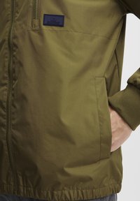 Olive green bomber jacket made of smooth nylon, featuring a front zipper, ribbed cuffs, and a label patch on the chest.