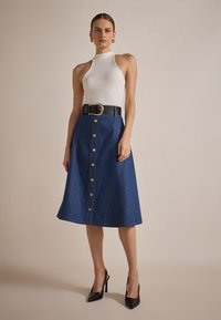 Jeansrok met knopen aan de voorkant, licht uitlopende ontwerpen, gecombineerd met een geribbelde witte haltertop en een zwarte riem met een gouden gesp. Zwarte hakken.