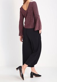 Top lungo maniche in glitter color bordeaux con scollatura a V profonda sul retro, abbinato a pantaloni neri larghi e scarpe con tacco nere. Design elegante e strutturato.