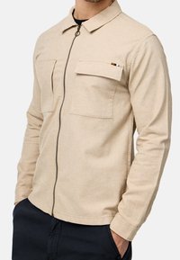 Veste beige à manches longues avec fermeture éclair à l’avant, deux poches poitrine, portée par-dessus une chemise noire, associée à un pantalon foncé.