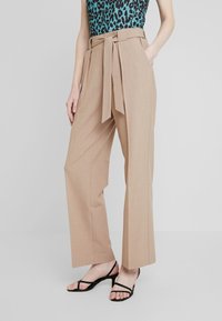 Beige wijde broek met een stoffen riem, scherpe plooien, zijzakken en een gladde textuur. Gecombineerd met zwarte sandaaltjes met bandjes.