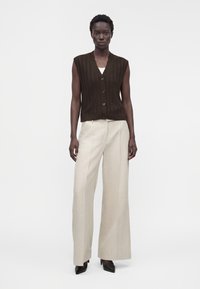 Femme portant un gilet marron sans manches boutonné sur un haut blanc, un pantalon crème à jambes larges et des talons noirs pointus, debout devant un fond uni.