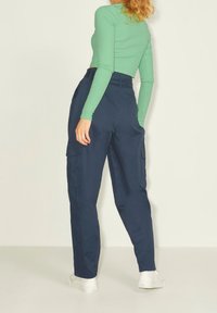 Kvinna som bär högmidjade marinblå cargobyxor med sidofickor, grön långärmad topp och vita sneakers, stående mot en enkel bakgrund.