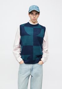 Πλεκτό αμάνικο πουλόβερ σε σκούρο μπλε και τιρκουάζ γεωμετρικό σχέδιο, φορεμένο πάνω από ένα ανοιχτό ροζ μακρυμάνικο πουκάμισο. Αξεσουάρ περιλαμβάνουν ένα τζιν καπέλο μπέιζμπολ.