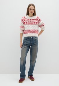 Maglione lavorato a maglia color crema con motivi geometrici rossi, abbinato a jeans blu slavati con leggere macchie e scarpe da ginnastica rosse.