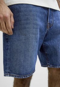 Shorts in denim di colore blu scuro con una trama liscia, caratterizzati da accenti di cucitura e un orlo dritto. Vista ravvicinata con la mano posizionata.