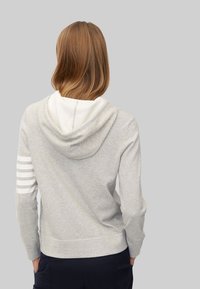 Šedá mikina s bílým lemováním kapuce, hladkou texturou a třemi bílými pruhy na levém rukávu.