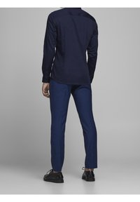 Camisa de botones azul marino con mangas largas, combinada con pantalones azul ajustados y zapatos negros, vista desde la parte de atrás. Tejido suave, diseño ajustado.