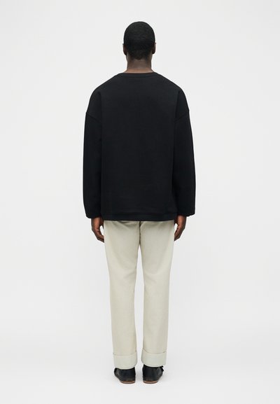 Άνδρας που στέκεται με την πλάτη, φοράει μαύρο πουκάμισο με μακριά μανίκια, ανοιχτόμπεζ παντελόνι με μανσέτες και μαύρα επίσημα παπούτσια σε απλό φόντο.