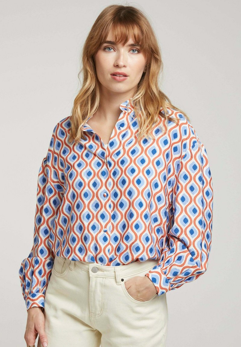 Chemise à motifs avec des formes géométriques bleues et oranges sur fond blanc, à coupe décontractée et manches longues.
