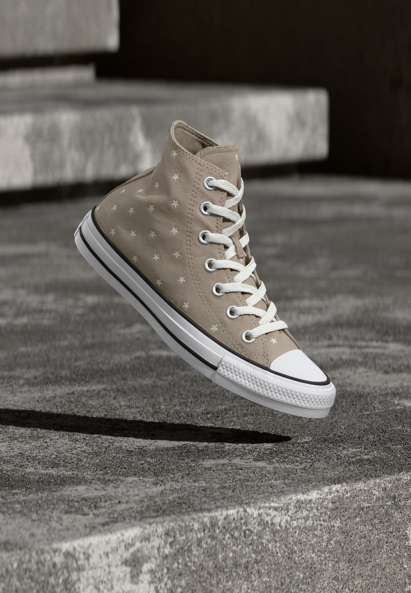 Beige høytliggende joggesko med hvitt stjernemønster og lisser, hengt over strukturerte betongtrinn som kaster en skygge under.