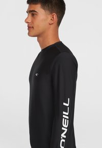 Musta pitkähihainen rash guard, jossa on pyöreä pääntie, sileä pinta ja valkoinen "O'Neill" -logo painettuna vasempaan hihaan.
