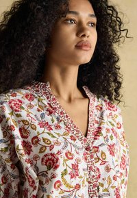 Bluzka w kwiatowy wzór w kolorze białym, ozdobiona czerwonymi i zielonymi motywami, z dekoltem w serek oraz haftowanymi akcentami wzdłuż dekoltu i guzików.