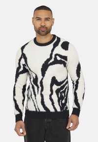Pull en tricot crème avec motif abstrait noir, doté d'un col rond et d'un tissu texturé avec une finition duveteuse.