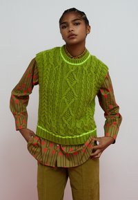 Giovane che indossa un gilet verde brillante lavorato a maglia sopra una camicia oliva con motivo a tulipani rossi, in piedi con le mani infilate nelle tasche dei pantaloni.