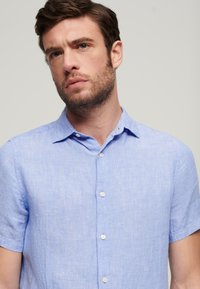 Camisa de manga corta azul claro, con botones, fabricada en una mezcla de lino texturizado, que presenta un cuello estándar y botones blancos en el frente.