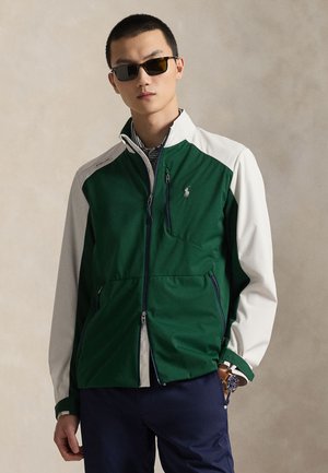 Άνδρας που φορά πράσινο και λευκό μπουφάν με φερμουάρ, μπλε παντελόνι, γυαλιά ηλίου και ρολόι χειρός, στέκεται μπροστά σε μια μπεζ επιφάνεια.