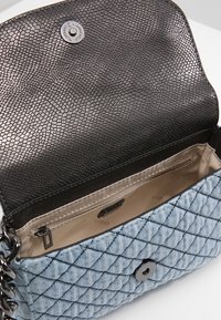 Borsa a tracolla in denim blu chiaro trapuntato con tracolla nera a catena, risvolto aperto che mostra un interno metallizzato e una fodera con tasca con zip beige.