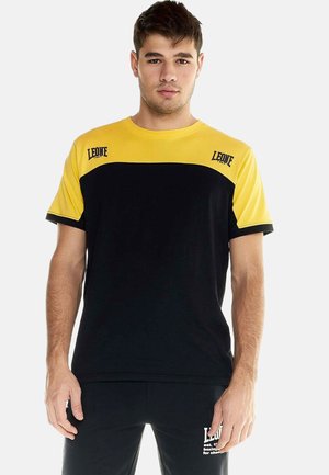 Jeune homme portant un t-shirt à manches courtes noir et jaune de la marque Leone et un pantalon noir, debout devant un fond clair uni.