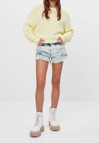 Gelber flauschiger Pullover, hellblaue, verwaschene Jeansshorts mit ausgefransten Kanten und weiße Plateau-Schnürstiefel mit durchsichtigen Sohlen.