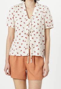 Blusa de manga corta en tejido crema con patrón de rosas rojas, con cuello tipo camisa y un lazo en la parte delantera. Combinada con pantalones cortos naranjas.