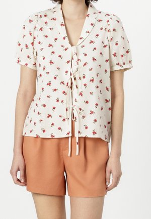 Blusa a maniche corte in tessuto crema con motivo a rose rosse, dotata di colletto e un nastro davanti. Abbinata a pantaloni corti arancioni.