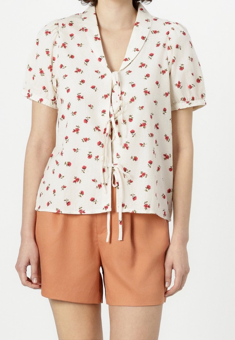 Blusa de manga corta en tejido crema con patrón de rosas rojas, con cuello tipo camisa y un lazo en la parte delantera. Combinada con pantalones cortos naranjas.