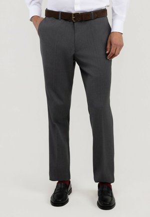 Hombre vestido con pantalones de vestir grises, cinturón marrón, camisa blanca, calcetines rojos y zapatos de vestir negros con las manos descansando cerca de los bolsillos.