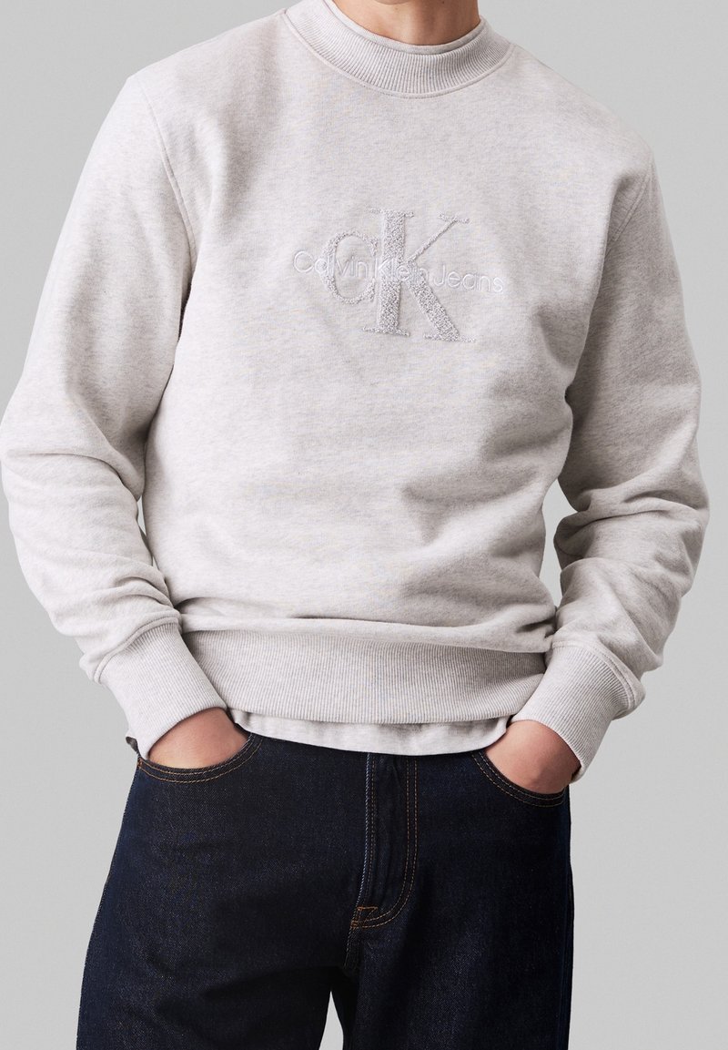 Sweat-shirt gris clair avec le logo "Calvin Klein Jeans" porté avec un jean bleu foncé, mains dans les poches, sur un fond gris uni.
