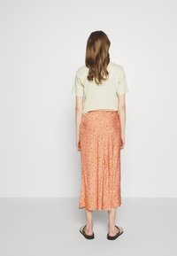 Vaaleanvihreä t-paita yhdistetty keskipitkään, liehuvaan oranssiin hameeseen, jossa on pieni kukkakuviointi. Malli käyttää mustia sandaaleja.