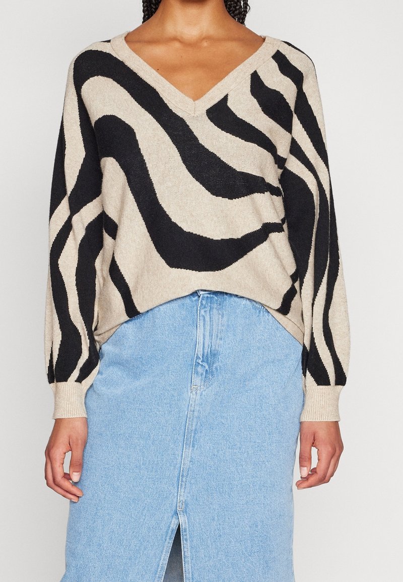 Femme portant un pull en V beige et noir à rayures abstraites, associé à une jupe en jean bleu clair avec une fente frontale.