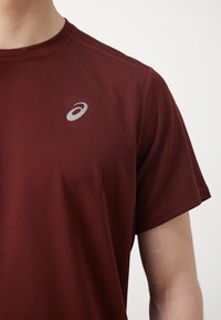 Burgunderfarget treningst-skjorte laget av lett materiale, har korte ermer, rund hals og et reflekterende logo på brystet.