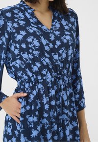 Sininen kukallinen mekko, jossa on V-pääntie, pitkät hihat ja joustava vyötärö, valmistettu kevyestä kankaasta, jossa on tumma sininen tausta.