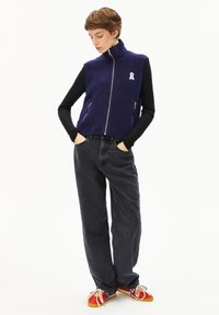 Lila Fleece-Weste mit hohem Kragen und Frontreißverschluss, mit zwei Reißverschlusstaschen und einem weißen Bandemblem. Kombiniert mit schwarzen Jeans und roten Schuhen.
