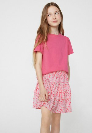 Młoda dziewczyna z długimi włosami, ubrana w jasnoróżowy t-shirt oraz różową kwiecistą warstwową spódnicę na jednolitym białym tle.