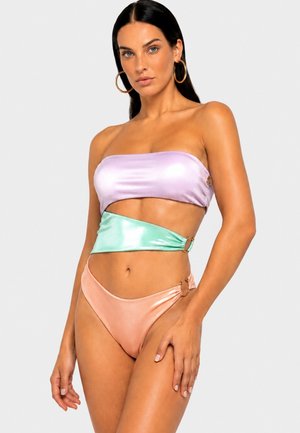 Donna con lunghi capelli scuri che indossa un bikini a blocchi di colore pastello con grandi orecchini a cerchio su uno sfondo chiaro uniforme.
