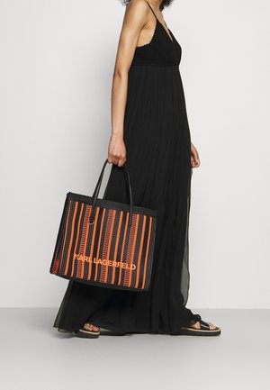 Femme portant une longue robe noire et des sandales noires, tenant un sac fourre-tout à rayures noires et orange de Karl Lagerfeld avec deux poignées.