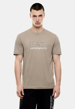 Giovane uomo che indossa una t-shirt beige EA7 Emporio Armani e pantaloni neri con il logo Emporio Armani sul lato, in piedi davanti a uno sfondo semplice.