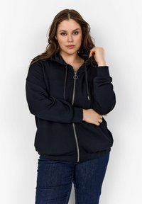 Sudadera negra con cremallera y cordón, de mangas largas y ribete en el dobladillo. Textura suave con una silueta ajustada, combinada con pantalones oscuros.