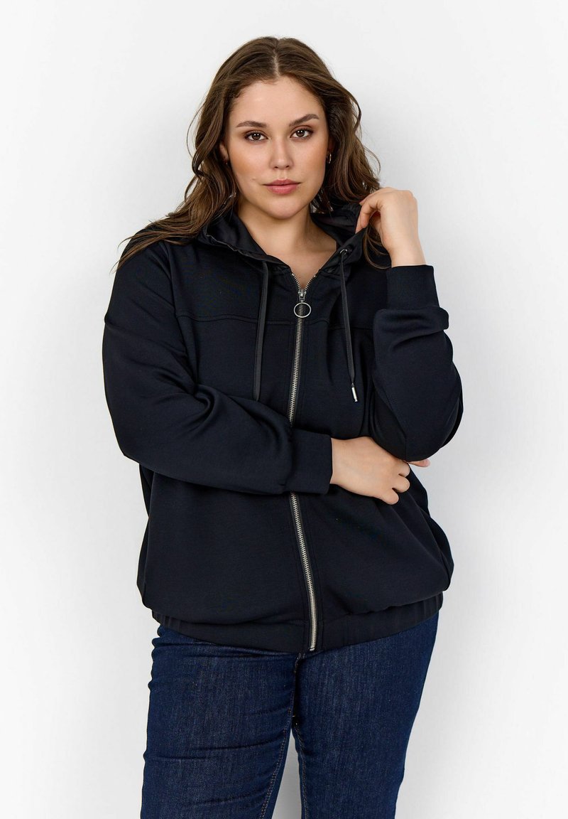 Sudadera negra con cremallera y cordón, de mangas largas y ribete en el dobladillo. Textura suave con una silueta ajustada, combinada con pantalones oscuros.