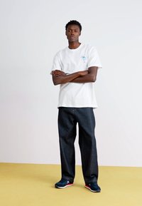 Λευκή βαμβακερή μπλούζα με γραφικό σχέδιο μπροστά, σκούρα μπλε τζιν παντελόνια σε φαρδιά γραμμή και ναυτικό αθλητικά παπούτσια με κόκκινες και λευκές λεπτομέρειες, σε ανοιχτό φόντο.