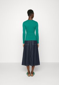 Mujer de pie de espaldas, vistiendo una camiseta verde ajustada de manga larga, una falda midi de mezclilla oscura con bolsillos traseros y sandalias verdes con suelas de corcho.