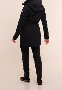 Zwarte waterdichte jas met capuchon, getailleerde taille en zijzakken met rits. Gecombineerd met zwarte broek en enkellaarsjes. Overal een gladde textuur.