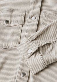 Camicia beige a righe con due tasche sul petto, bottoni a pressione argentati e texture morbida. Presenta dettagli ai polsini e cuciture pulite.