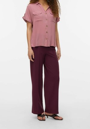 Femme portant une chemise à manches courtes rose poussiéreux à boutons, un pantalon large violet foncé et des sandales à talons ouvertes noires.