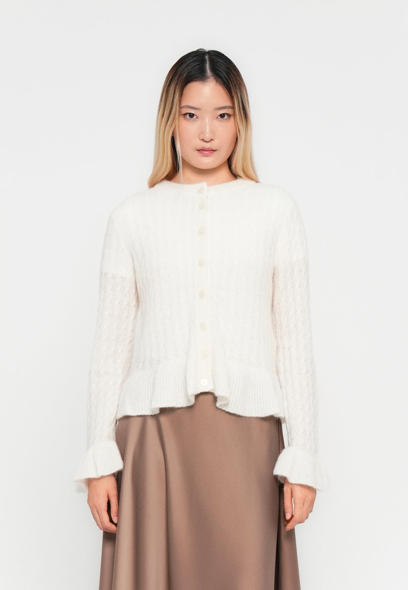 Junge Frau mit glattem, ombrefarbenem Haar, die einen weißen, knöpfbaren Strickpullover mit Rüschensäumen und einen braunen SatRock trägt.