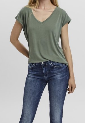 Frau trägt olivgrünes V-Ausschnitt-Top mit Kappenärmeln und eng anliegende blaue Jeans, steht mit einer Hand in der Hüfte vor einem schlichten Hintergrund.